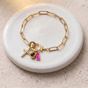 A gold paperclip chain bracelet with three charms, including a pink rectangular stone, a black oval stone, and a small gold cross, displayed on a white ceramic dish over a textured beige fabric background.