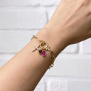 A gold chain bracelet with multiple charms, including a cross, a small black bead, and a rectangular magenta gemstone, shown worn on a person's wrist against a white brick wall background.