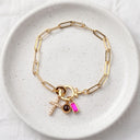 A gold paperclip chain bracelet with three charms, including a gold cross with rhinestones, a dark brown oval stone, and a bright pink rectangular stone, displayed on a speckled white ceramic dish.