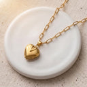 Gold heart locket closed on a paperclip chain, resting on a white ceramic dish
