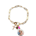 A gold paperclip chain bracelet with a clasp featuring a small gold cross, a pink rectangular stone, a dark round stone, and a circular custom photo charm showing a family portrait, set against a plain white background.